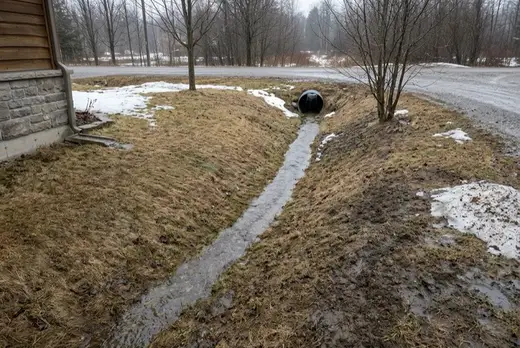Meltwater running along a grassed swale beside a foundation and into a clear culvert at the roadside