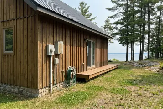The summer side of a timber chalet showing the exterior tap, hose reel and hydro meter, with Georgian Bay through the pines